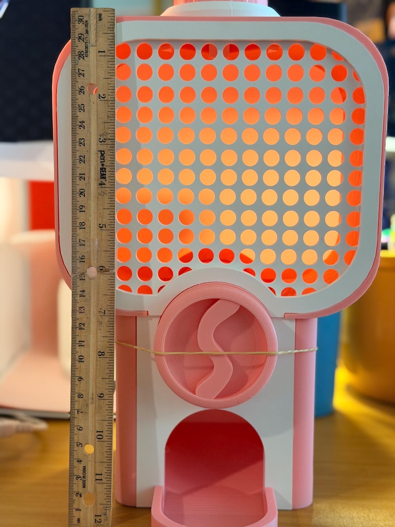 May include: A pink and white plastic gumball machine featuring a grid-patterned front panel and a circular dispensing mechanism. A wooden ruler is placed vertically along the left side, showing the machine stands approximately 29 cm tall.