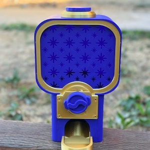 May include: A blue and gold gumball machine featuring a square-shaped container with a star pattern on the front panel. The device includes a circular top lid, a central dispensing knob, and a small gold tray at the bottom for collecting items.