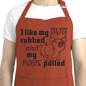 May include: Rust-colored apron with black text and a pig illustration. The text reads "I like my BUTT rubbed and my PORK pulled." The pig illustration shows a smiling pig from the rear.