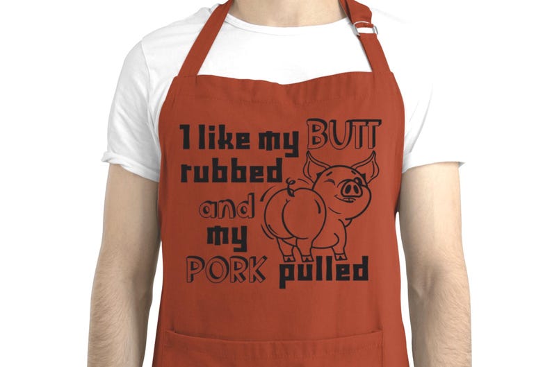 May include: Rust-colored apron with black text and a pig illustration. The text reads "I like my BUTT rubbed and my PORK pulled." The pig illustration shows a smiling pig from the rear.