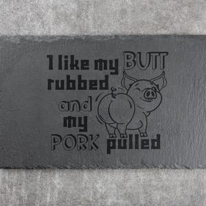 May include: A rectangular, dark gray slate serving board with the text "I like my butt rubbed and my pork pulled" and a pig illustration. A red and black checkered cloth and grilling utensils are on either side.