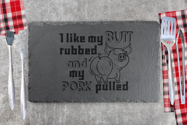 May include: A rectangular, dark gray slate serving board with the text "I like my butt rubbed and my pork pulled" and a pig illustration. A red and black checkered cloth and grilling utensils are on either side.