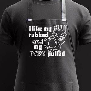 May include: Black apron with white text and a pig illustration. The text reads "I like my butt rubbed and my pork pulled." The pig illustration is a cartoon-style drawing. The apron has a pocket and adjustable neck strap.