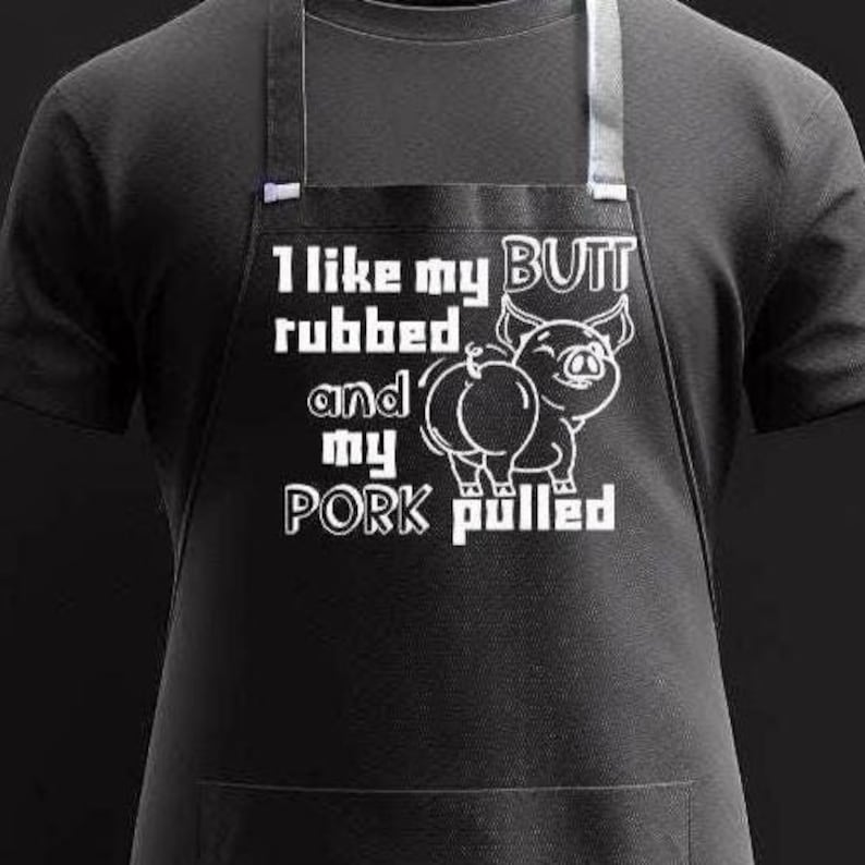 May include: Black apron with white text and a pig illustration. The text reads "I like my butt rubbed and my pork pulled." The pig illustration is a cartoon-style drawing. The apron has a pocket and adjustable neck strap.