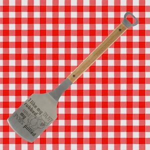 May include: A stainless steel spatula with a wooden handle and bottle opener. The spatula features the text "I like my butt rubbed and my pork pulled" with a pig graphic. The background is a red and white checkered pattern.