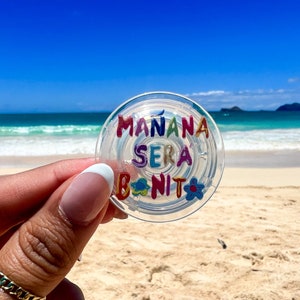 May include: A clear, circular phone grip with the words "MAÑANA SERA BONITO" in colorful, raised lettering. The grip features flower and leaf designs. The background is a beach scene with blue sky and ocean.