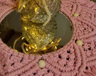 Könnte beinhalten: Ein rosa Makramee-Wandbehang mit Holzperlen und einem runden Spiegel in der Mitte. Der Spiegel spiegelt eine Glasvase mit Lichtern darin wider.