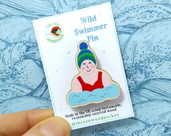 May include: Wooden pin of a person wearing a blue, green and white hat swimming in blue water. The pin is made in the UK using sustainable, responsibly sourced wood. The text "Wild Swimmer Pin" and "@thezenwoodpecker" are on the card.