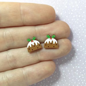May include: Two miniature Christmas pudding earrings. Each earring is brown with white icing, small brown dots, and a green holly leaf and red berry accent. The earrings are small and festive.