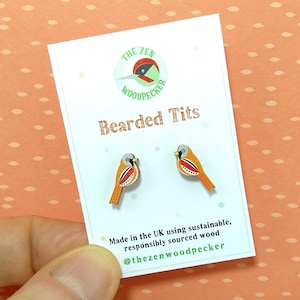 Könnte beinhalten: Holzohrstecker mit Vogeldesign. Die Ohrringe sind orange, rot und grau, mit einem schwarzen Schnabel. Die Ohrringe befinden sich auf einer weißen Karte mit dem Text "Bearded Tits" und dem Logo von "The Zen Woodpecker".
