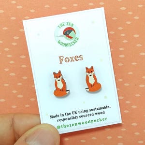 以下が含まれることがあります： 白いカードに、キツネの形をしたイヤリングのペア。イヤリングはオレンジと白で、キツネのデザインです。カードには「Foxes」の文字とロゴがあります。カードには「Made in the UK using sustainable, responsibly sourced wood」とも書かれています。