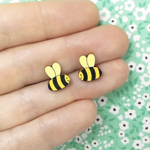 Peut inclure: Une paire de boucles d'oreilles en bois en forme d'abeille. Les abeilles sont jaunes et noires avec un visage souriant. Les boucles d'oreilles sont petites et délicates.