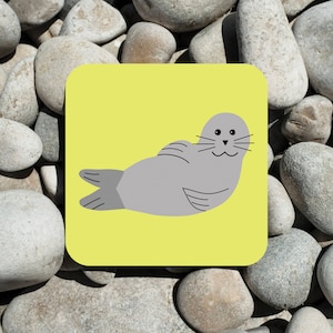Può includere: Una foca grigia con occhi neri e un naso nero è sdraiata a pancia in giù con le pinne distese. La foca è su uno sfondo giallo.
