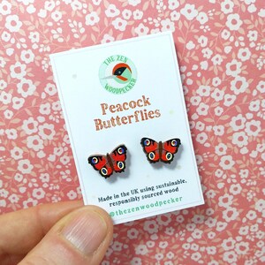 Puede incluir: Un par de pendientes de madera con forma de mariposa pavo real. Los pendientes son rojos, negros y blancos con un fondo marrón. Los pendientes están hechos en el Reino Unido con madera sostenible y de origen responsable. El texto en la tarjeta dice "The Zen Woodpecker Peacock Butterflies Made in the UK using sustainable, responsibly sourced wood @thezenwoodpecker"