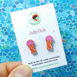Puede incluir: Un par de pendientes de madera de color rosa y naranja con forma de medusa. Los pendientes están sobre una tarjeta blanca con el texto "THE ZEN WOODPECKER" y "Jellyfish". La tarjeta también dice "Hecho en el Reino Unido con madera sostenible y de origen responsable" y "@thezenwoodpecker".