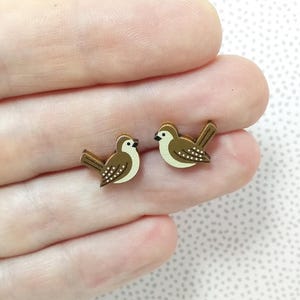 May include: A pair of brown wooden earrings shaped like birds with white accents. The birds have brown eyes and are facing to the right.