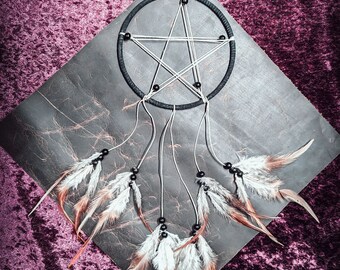 May include: A black dreamcatcher with a white pentagram and black beads hanging from a gray background. The dreamcatcher has brown and white feathers.