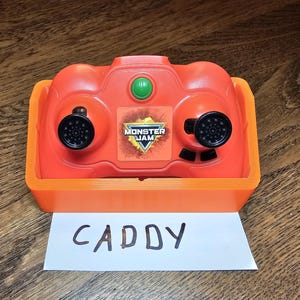 May include: Orange and black Monster Jam remote control with a green button and the words "Monster Jam" on the front. The remote control is sitting on a white piece of paper with the word "CADDY" written on it.