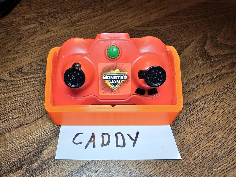 May include: Orange and black Monster Jam remote control with a green button and the words "Monster Jam" on the front. The remote control is sitting on a white piece of paper with the word "CADDY" written on it.