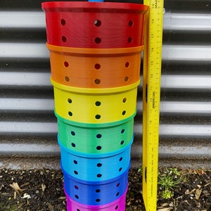 May include: A stack of six colorful plastic containers with holes. The containers are red, orange, yellow, green, blue, and purple. The containers are stacked on top of each other and are about 18 inches tall.