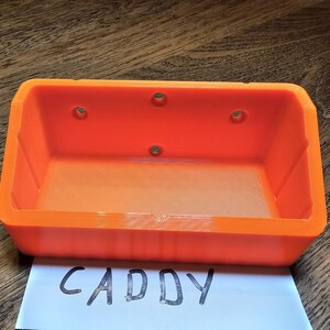 May include: An orange plastic caddy with four holes on the top and the word "CADDY" written on a white piece of paper below it.