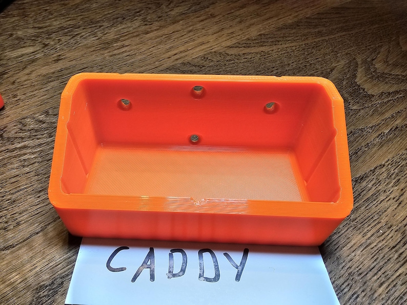 May include: An orange plastic caddy with four holes on the top and the word "CADDY" written on a white piece of paper below it.