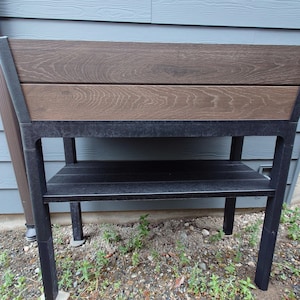 May include: A rectangular outdoor planter with a dark brown wood-look top and a black metal frame. The planter has a lower shelf and is set against a blue-gray siding background. The planter is designed for gardening.