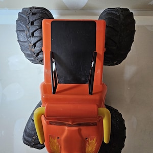 May include: Orange toy monster truck with black tires and yellow handlebars. The truck has a black roof and flames on the sides.