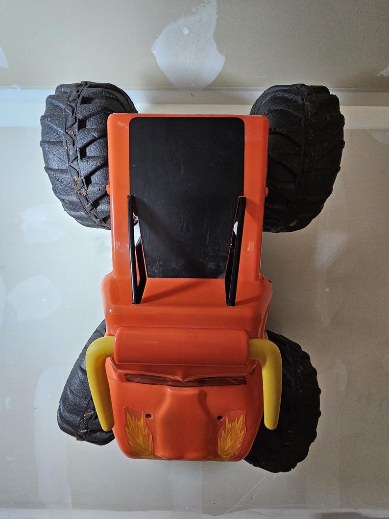 May include: Orange toy monster truck with black tires and yellow handlebars. The truck has a black roof and flames on the sides.
