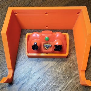 May include: Orange plastic wall mount for a Monster Jam remote control. The mount has two hooks on the bottom and a slot for the remote control.