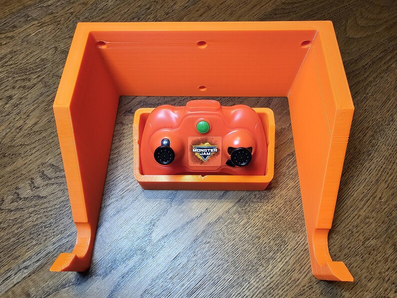 May include: Orange plastic wall mount for a Monster Jam remote control. The mount has two hooks on the bottom and a slot for the remote control.