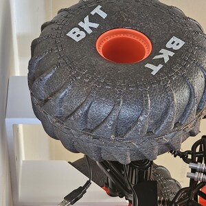 May include: A black toy monster truck with large black tires that say "BKT" on the side. The truck is on a white shelf with a black USB cable plugged into the truck.