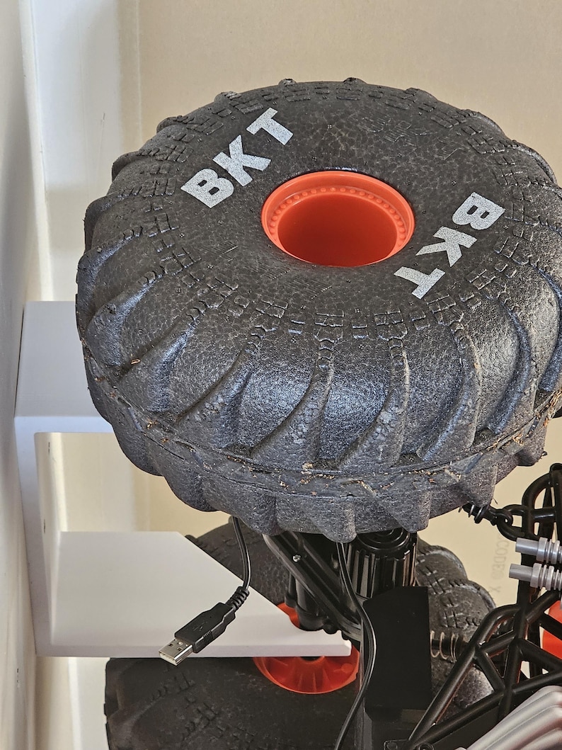 May include: A black toy monster truck with large black tires that say "BKT" on the side. The truck is on a white shelf with a black USB cable plugged into the truck.