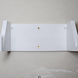 May include: White wall mount shelf with two screw holes. The shelf has two side supports that are angled to hold an object.