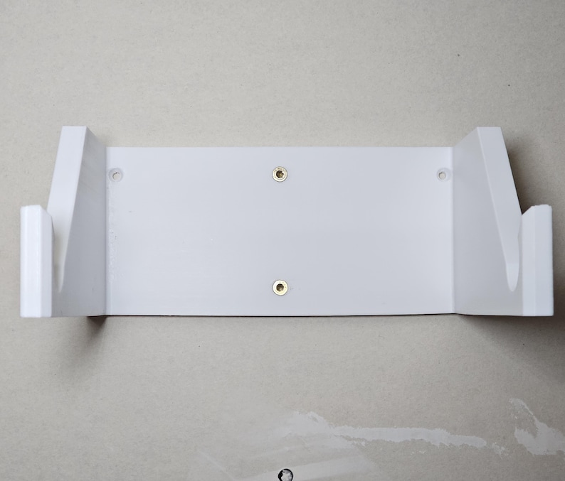 May include: White wall mount shelf with two screw holes. The shelf has two side supports that are angled to hold an object.
