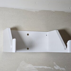 May include: White wall-mounted shelf with two angled supports for holding items. The shelf is made of plastic and has two screw holes on the front.