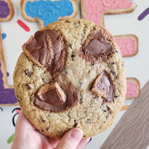 Peut inclure: Un gros cookie aux p&eacute;pites de chocolat avec quatre morceaux de chocolat au lait sur le dessus. Le cookie est de couleur brun clair et a une texture l&eacute;g&egrave;rement croustillante.