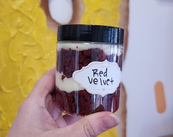 May include: A glass jar filled with layers of red velvet cake and white frosting. The jar has a black lid and a white label that says "Red Velvet".