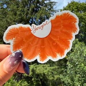 May include: A die-cut sticker featuring a stylized orange feather design with a white border. The sticker has the word "Showgirl" in red script. The sticker is held in front of a green leafy background.