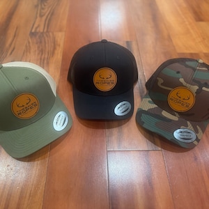 May include: Three baseball caps in olive green, black, and camouflage patterns. Each hat features a leather patch with the text "TIMESAVER ROPES" and a deer antler design. The hats have a curved brim and a mesh back.