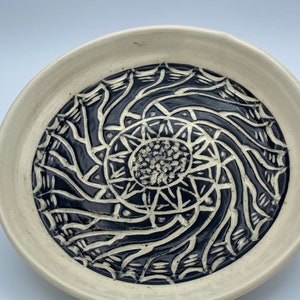 May include: A handmade ceramic bowl with a cream-coloured rim and a dark blue, intricate spiral design in the centre. The design features a central starburst pattern surrounded by swirling lines and shapes.