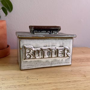 May include: A ceramic butter dish with a rectangular shape and a lid. The word "BUTTER" is embossed on the front. The dish has a white and gray glaze with brown accents on the lid.