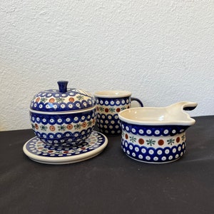 May include: A set of Polish pottery with a blue and white floral pattern. The set includes a lidded apple-shaped container on a saucer, a mug, and a small creamer. The pottery has a hand-painted design with red, green, and brown accents.