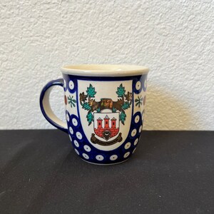 Puede incluir: Una taza de cer&aacute;mica con un dise&ntilde;o floral azul y blanco. La taza presenta un escudo detallado con un edificio rojo y blanco, follaje verde y el texto "750" y "Bolesławiec". El asa es azul.