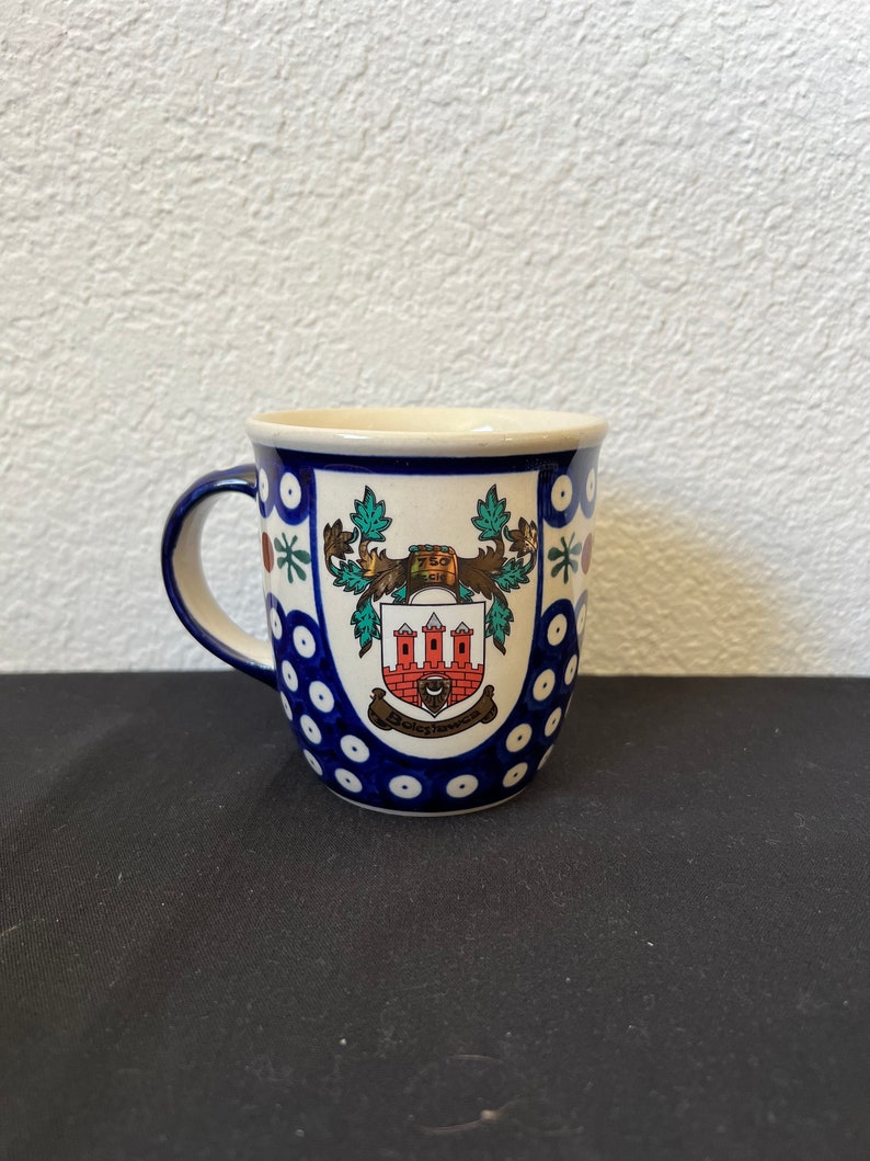 Puede incluir: Una taza de cer&aacute;mica con un dise&ntilde;o floral azul y blanco. La taza presenta un escudo detallado con un edificio rojo y blanco, follaje verde y el texto "750" y "Bolesławiec". El asa es azul.