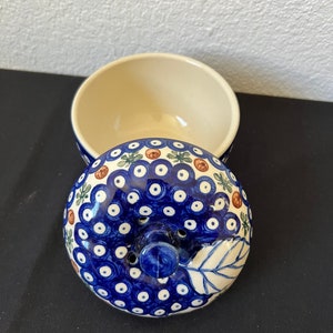 Puede incluir: Un cuenco de cer&aacute;mica con tapa. La tapa est&aacute; decorada con un patr&oacute;n azul y blanco de puntos y detalles florales. El interior del cuenco es de color crema y la tapa tiene un peque&ntilde;o asa. El cuenco est&aacute; abierto, revelando su interior.