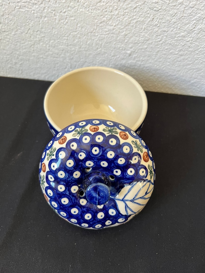Puede incluir: Un cuenco de cer&aacute;mica con tapa. La tapa est&aacute; decorada con un patr&oacute;n azul y blanco de puntos y detalles florales. El interior del cuenco es de color crema y la tapa tiene un peque&ntilde;o asa. El cuenco est&aacute; abierto, revelando su interior.