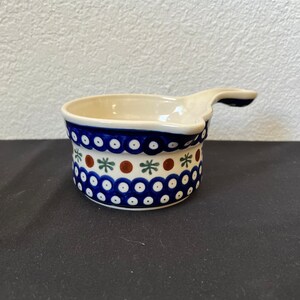 Puede incluir: Un peque&ntilde;o cuenco de cer&aacute;mica color crema con asa. El cuenco presenta un patr&oacute;n de puntos azules y blancos con detalles en marr&oacute;n y verde. El borde es azul oscuro liso. El asa tambi&eacute;n es azul.