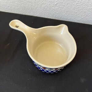 Puede incluir: Salsera de cer&aacute;mica color crema con asa y un estampado floral azul y blanco alrededor del borde. El asa tiene un peque&ntilde;o agujero. El plato est&aacute; vac&iacute;o y sobre una superficie oscura.