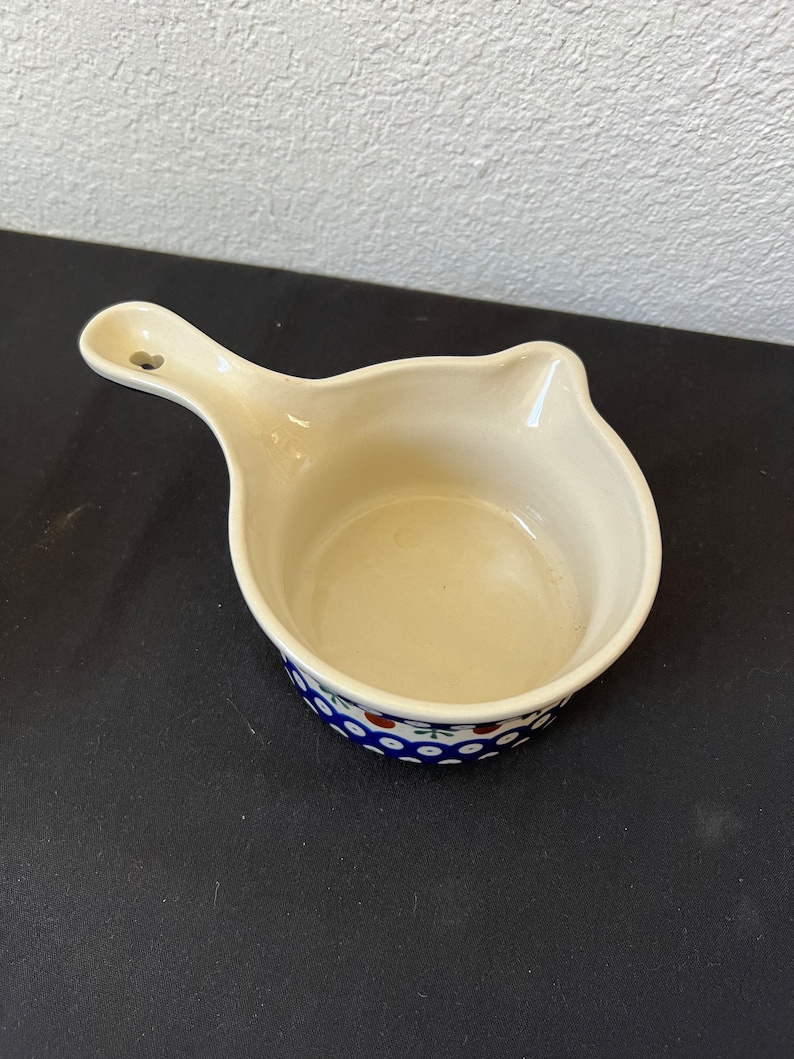 Puede incluir: Salsera de cer&aacute;mica color crema con asa y un estampado floral azul y blanco alrededor del borde. El asa tiene un peque&ntilde;o agujero. El plato est&aacute; vac&iacute;o y sobre una superficie oscura.